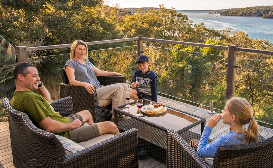 2 adults and 2 kids lounging on the balcony at Hilltop Cottage looking over the water in Royal National Park. Credit: John Spencer © DCCEEW