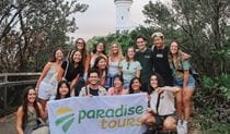 Group of people standing behind Paradise Tours flag with Cape Byron Lighthouse in the background. Photo: Nerea Sánchez &copy; Destination NSW