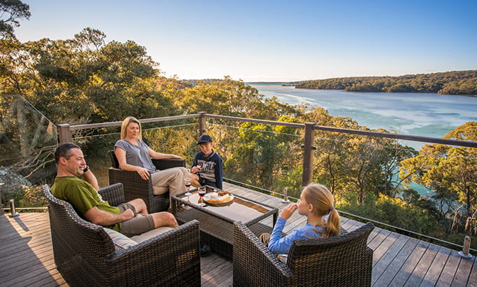 2 adults and 2 kids lounging on the balcony at Hilltop Cottage looking over the water in Royal National Park. Credit: John Spencer &copy; DCCEEW