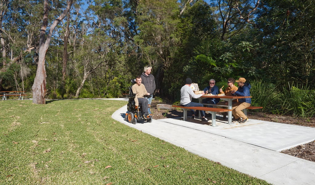 People share lunch at a picnic table, as a man in a wheelchair and his friend pass by, Somersby Falls picnic area, Brisbane Water National Park. Credit: Stuart Cohen, Bottle Brush Media © DCCEEW