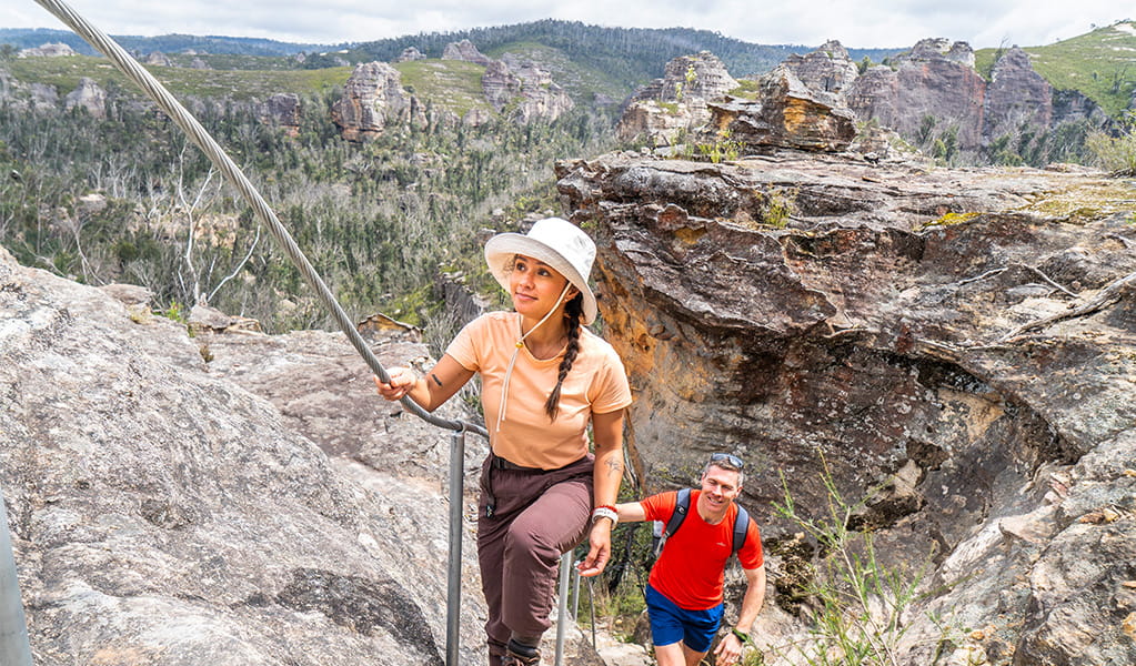 2 people holding onto a cable while climbing a steep section of Miners Pass link track, an optional Grade 4 section that connects with Lost City Walking track. Credit: Simone Cottrell &copy; DCCEEW