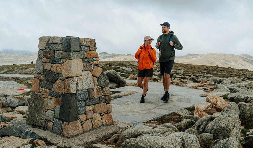 A man and woman walking towards the stone trig at the summit of Mount Kosciuszko with mountain peaks in the background. Credit: Remy Brand / DCCEEW &copy; Caravel Content