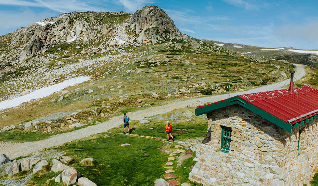 Seamans Hut in the foreground with 2 hikers walking towards the Snowy Mountain peaks of Kosciuszko National Park in the background. Credit: Daniel Parsons / DCCEEW &copy; Caravel Content