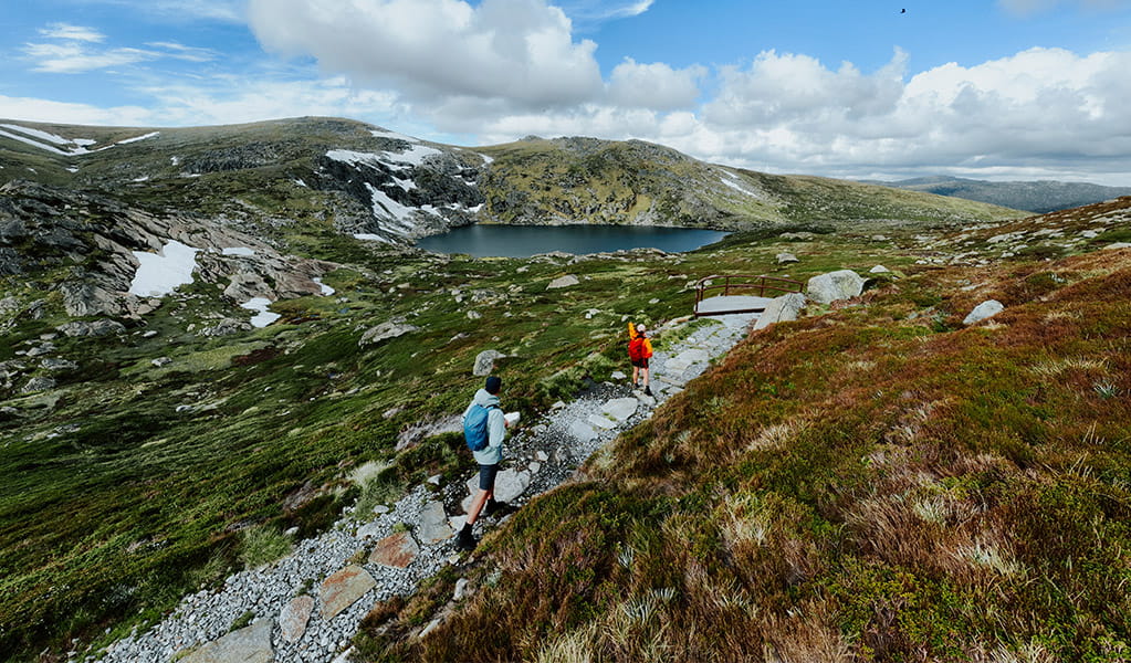 2 hikers walking on a gravel path on day 2 of Snowies Alpine Walk with Blue Lake and snow capped mountains in the background. Credit: Daniel Parsons / DCCEEW &copy; Caravel Content