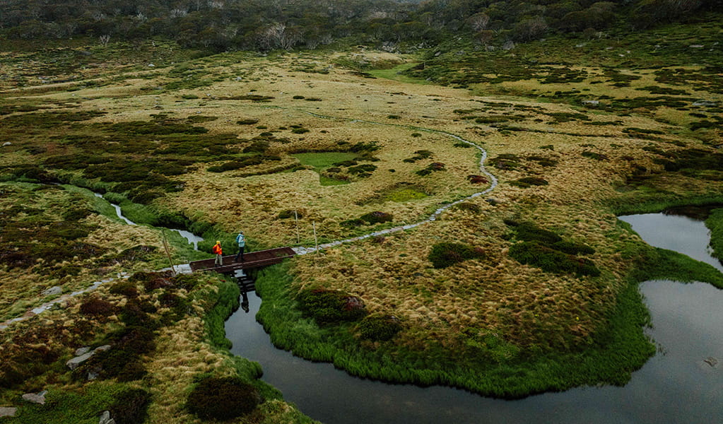 An aerial view of 2 hikers crossing a winding creek surrounded by marsh on Snowies Alpine Walk, Kosciuszko National Park. Credit: Daniel Parsons / DCCEEW &copy; Caravel Content