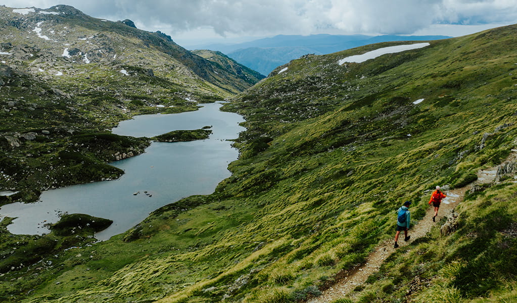 2 hikers on a track along Snowies Alpine Walk with a view of snow-capped mountains and a lake in the background. Credit: Daniel Parsons / DCCEEW &copy; Caravel Content