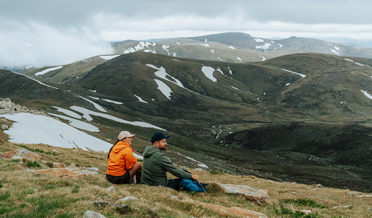 2 hikers sitting at the summit of Mount Kosciuszko looking at expansive views of the surrounding Snowy Mountains. Credit: Remy Brand / DCCEEW © Caravel Content