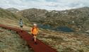 2 hikers walking on a raised path on day 2 of Snowies Alpine Walk with snow capped mountains in the background. Credit: Remy Brand / DCCEEW &copy; Caravel Content