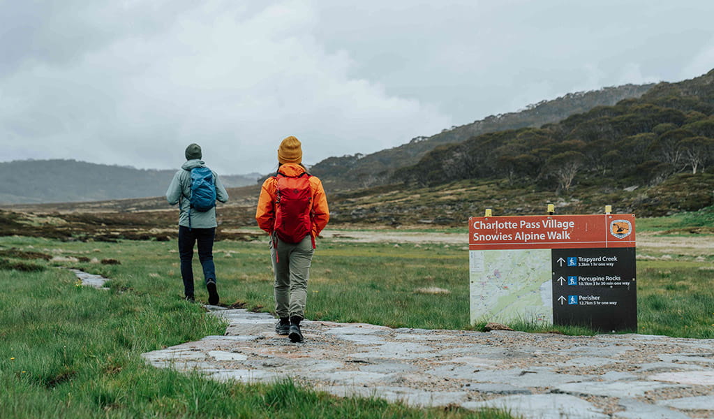 2 hikers walking past the track head sign at Charlotte Pass Village to Perisher Valley walk. Credit: Remy Brand / DCCEEW © Caravel Content