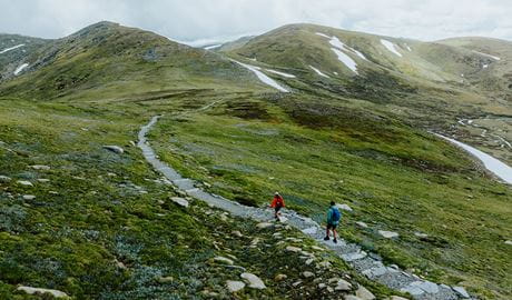 2 hikers on a track along Main Range walk with a view of snow-capped mountains and a lake in the background. Credit: Daniel Parsons / DCCEEW &copy; Caravel Content