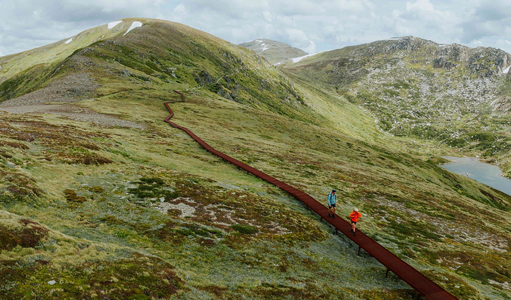 2 hikers on a raised track along Main Range walk with the Snowy Mountains in the background. Credit: Daniel Parsons / DCCEEW &copy; Caravel Content