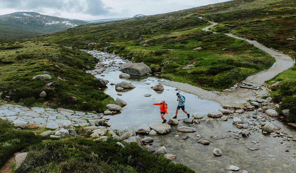 An aerial image of 2 people crossing the Snowy River by rock hopping along Main Range walk. Credit: Daniel Parsons / DCCEEW &copy; Caravel Content