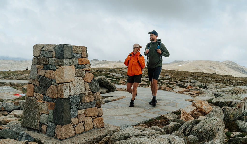 A man and woman walking towards the stone trig at the summit of Mount Kosciuszko with mountain peaks in the background. Credit: Remy Brand / DCCEEW &copy; Caravel Content