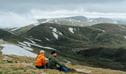 2 hikers sitting at the summit of Mount Kosciuszko admiring a view of snowy mountain peaks. Credit: Remy Brand / DCCEEW &copy; Caravel Content