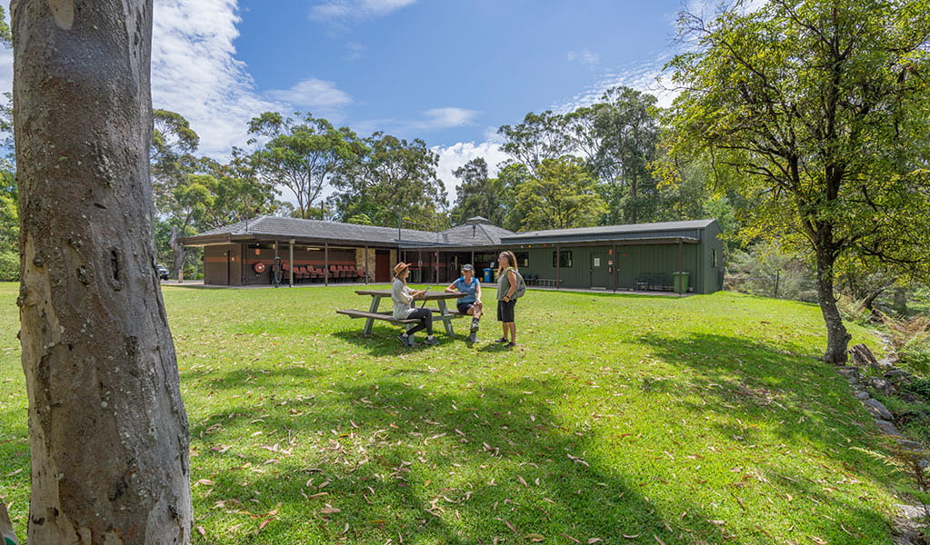 Two people sit on a picnic table bench and one person stands on grassy lawn behind Kalkari Discovery Centre. Photo: Johny Spencer &copy; DCCEEW