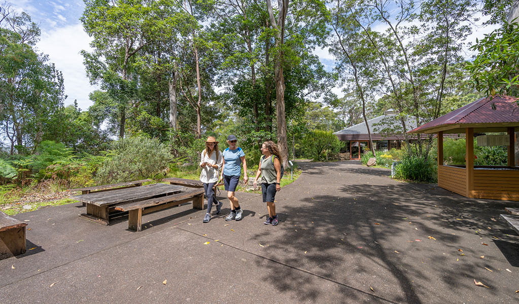 Three women walk along a flat concrete path behind Kalkari Discovery Centre, in Ku-ring-gai Chase National Park. Photo: Johny Spencer &copy; DCCEEW
