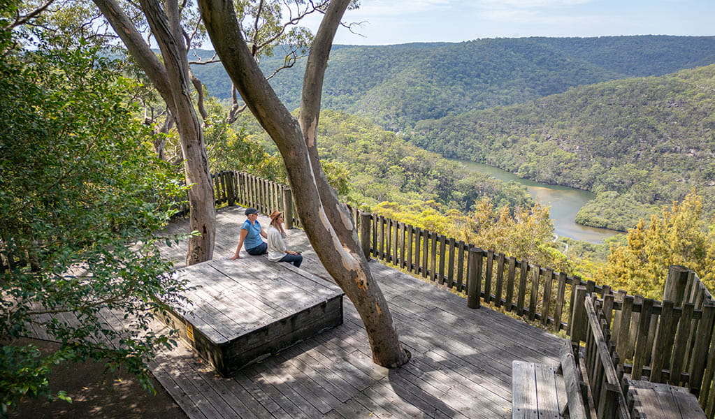 Two women sit on a timber bench while enjoying views at Kalkari lookout in Ku-ring-gai Chase National Park. Photo: Johny Spencer &copy; DCCEEW
