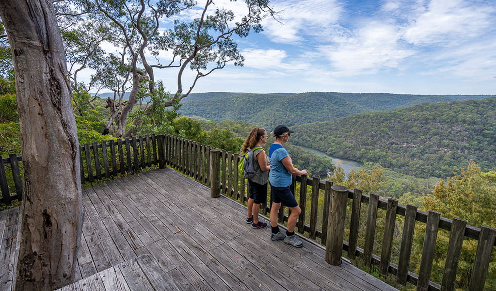 Two people stand at a timber fence atop Kalkari lookout timber platform, overlooking Cowan Creek and a forested valley in Ku-ring-gai Chase National Park. Photo: Johny Spencer &copy; DCCEEW