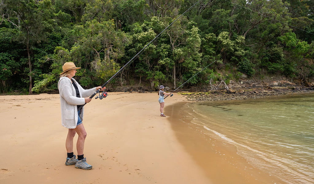 2 people beach fishing in The Basin lagoon with Ku-ring-gai Chase National Park greenery in the background. Credit: John Spencer/DCCEEW  &copy; DCCEEW