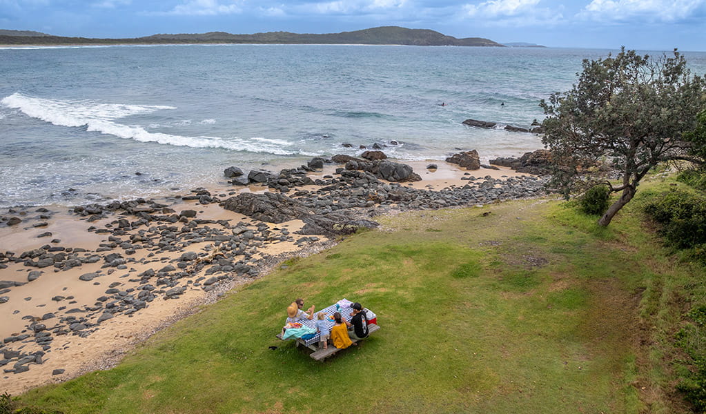 A family sitting at a picnic table by the beach at Point Plomer campground in Limeburners Creek National Park. Credit: John Spencer &copy; DCCEEW