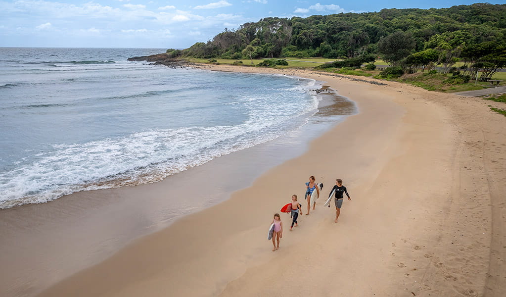 A family walking the beach with surfboards under their arms, next to Point Plomer campground in Limeburners Creek National Park. Credit: John Spencer &copy; DCCEEW