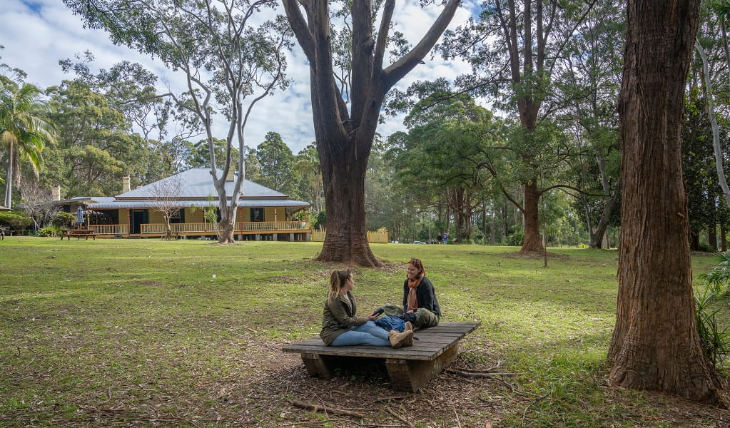 Two women have a relaxed chat sitting on a low wooden platform in the shade of gum trees, with Roto House in the background, Roto House Historic Site. Credit: John Spencer &copy; DCCEEW 