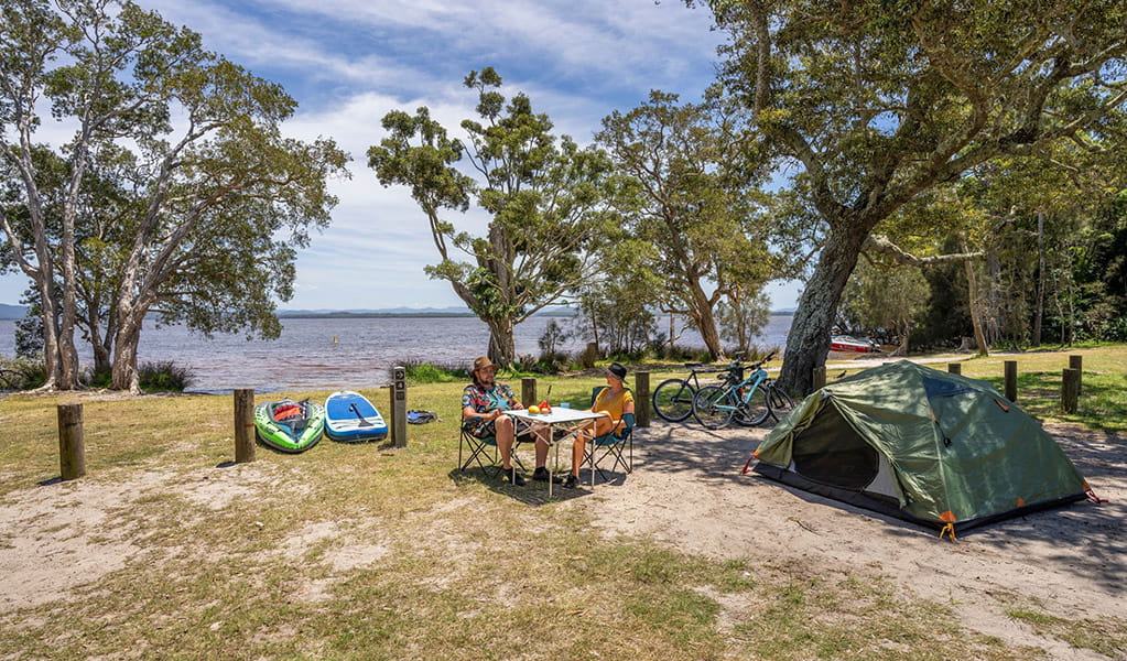 2 campers at a camping table near a tent by the water at Mungo Brush campground, Myall Lakes National Park. Credit: John Spencer/DCCEEW &copy; DCCEEW