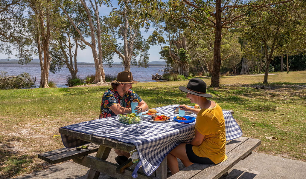 2 people having a picnic on a table by the water at Mungo Brush campground, Myall Lakes National Park. Credit: John Spencer/DCCEEW &copy; DCCEEW