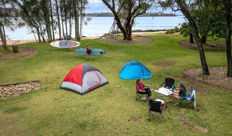 Campers sitting next to 2 tents at Bonnie Vale campground with 2 people walking towards the water with paddleboards. Credit: John Spencer/DCCEEW  &copy; DCCEEW