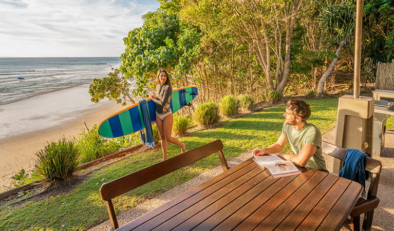 Woman holding a surfboard by the ocean in the backyard of Mildenhall cottage. Photo: DPIE/John Spencer