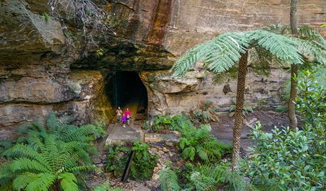 An image of Glow Worm Tunnel exit carved into a large rock wall with a family walking out to Penrose Gorge. Credit: John Spencer &copy; DCCEEW  